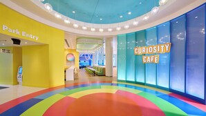 A brightly lit indoor space with a colorful floor design, a 'Park Entry' sign, and a 'Curiosity Café' sign. - Universal Kids Resort Hotel (Frisco)