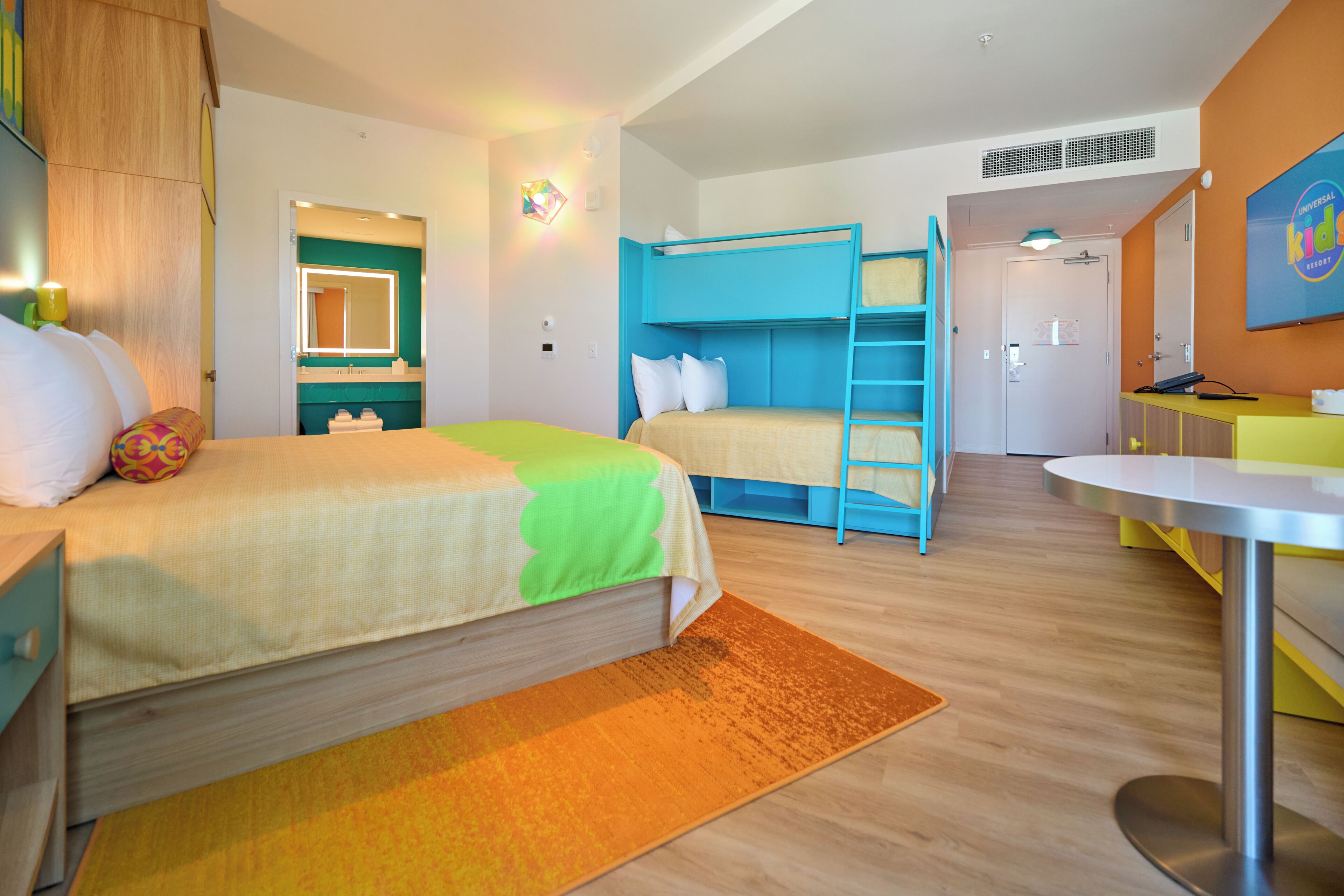 Habitación de hotel con literas, escritorio, silla, mesita pequeña, espejo y puerta de baño.