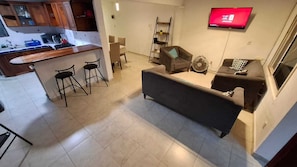 A modern living area with a sofa, a TV mounted on the wall, and a kitchen area with wooden cabinets and a bar stool. - Spacious, centrally located residence with a private pool! Perfect for Families! (Cancún)