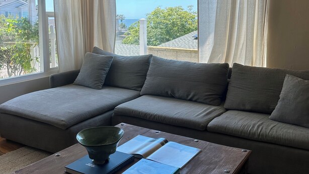 Ein modernes Wohnzimmer mit einem grauen Ecksofa, einem Couchtisch aus Holz mit einem Buch und einer grünen Schale, sowie großen Fenstern mit transparenten Vorhängen.