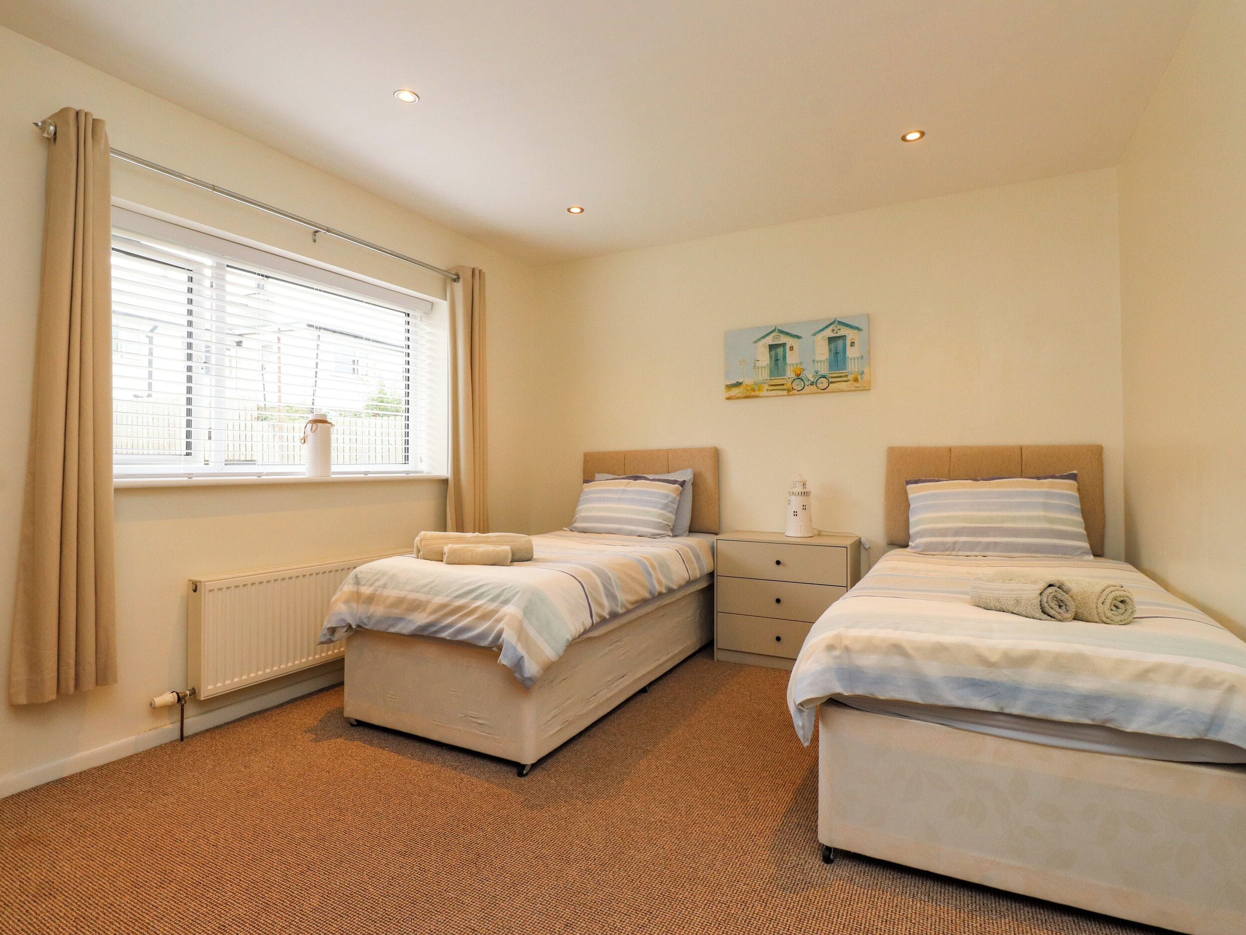 A bedroom with two beds, a radiator, a window with curtains, a small bedside table, and a painting on the wall.