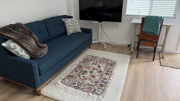 A living room with a dark blue sofa, a flat-screen TV, a patterned rug, a wooden chair, and a window with blinds. - Beautiful ADU with kitchen, washer and dryer. Near cafes, grocery stores and gym (Pasadena)