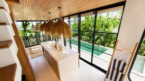 A modern kitchen with a large island, a dining area with a striped chair, and a view of a swimming pool through large windows. - 2BR Private Pool Villa • Near Cemagi Beach & Canggu (Badung)
