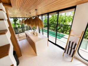 Une cuisine moderne dotée d’un grand îlot central, d’un coin repas avec une chaise à rayures et d’une vue sur une piscine, visible à travers de grandes fenêtres.