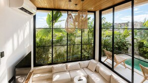 A modern living room with a large sectional sofa, a small round table, and a view of a pool and greenery through large windows. - 2BR Private Pool Villa • Near Cemagi Beach & Canggu (Badung)