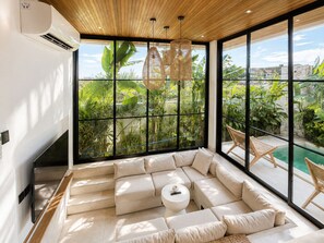 Un salon moderne doté d’un grand canapé d’angle, d’une petite table ronde et offrant une vue sur une piscine et un espace verdoyant grâce à de grandes fenêtres.