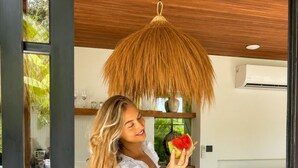 A person in a white dress holding a slice of watermelon, standing in a room with a wooden ceiling, a hanging light fixture, and a pool visible through a glass door. - 2BR Private Pool Villa • Near Cemagi Beach & Canggu (Badung)