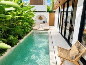 Un espace piscine moderne avec une piscine à eau claire, entouré d’une végétation luxuriante et agrémenté d’un fauteuil de relaxation en osier.