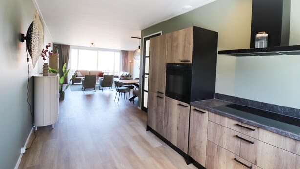 A modern kitchen with wooden cabinets, a black countertop, and a dining area in the background. - New, Luxury Holiday Home in the Heart of Friesland (Leons)