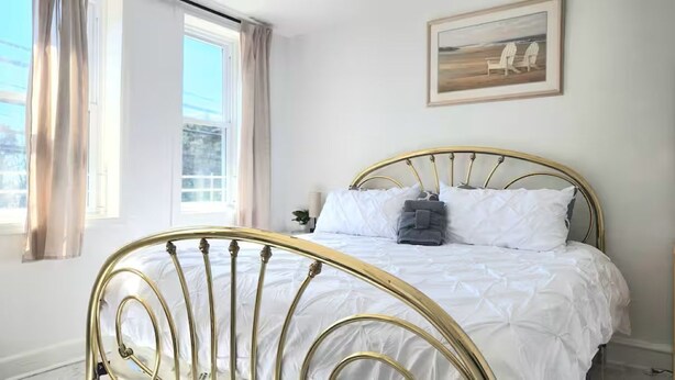 Un lit avec un cadre doré, une literie blanche et un tableau représentant des chaises accroché au mur.