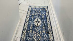 Un couloir doté d’un sol en marbre, d’un tapis bleu à motifs et d’une porte donnant sur une pièce où se trouvent un lit et une chaise.