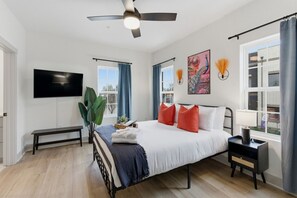 A bedroom with a bed, bedside table, lamp, TV, and large windows with curtains. - Boutique Loft w/ Patio, Rooftop Views + In-Unit W/D (Murfreesboro)