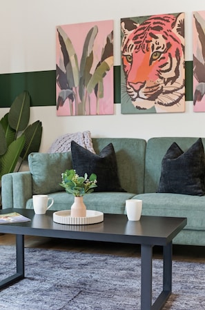 Un canapé vert avec deux coussins noirs, une table basse blanche avec une plante, et deux tasses blanches.