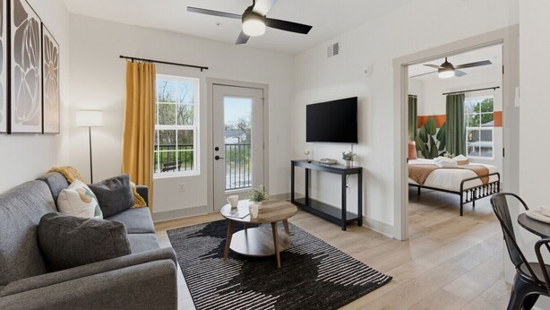 Uma sala moderna com sofá, mesa de centro e televisão. Há uma varanda com vista, e um quarto com cama e janela.