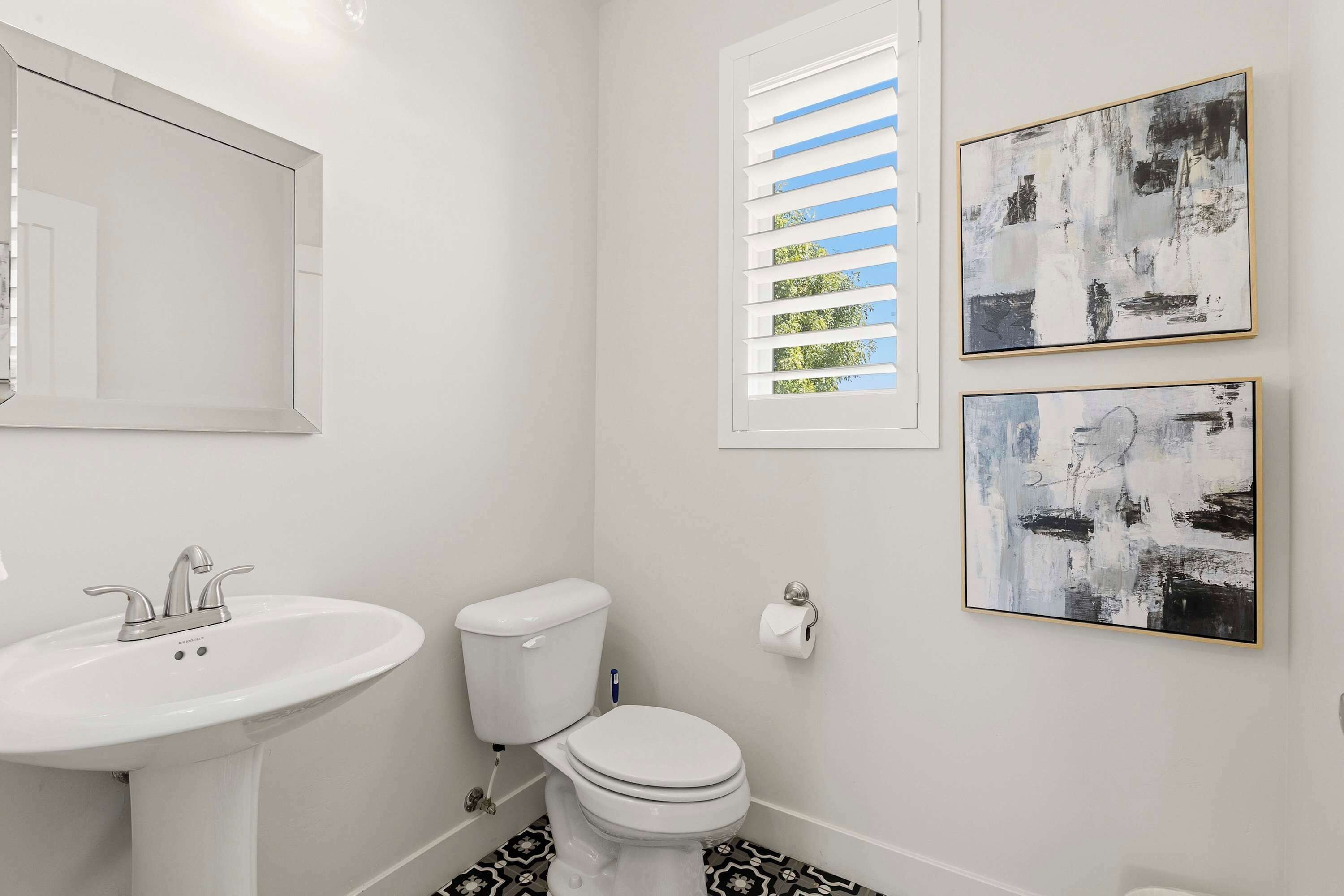 A bathroom with a white sink, toilet, and window with blinds. There are two abstract paintings on the wall.