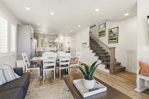 Una cocina moderna con armarios blancos, un comedor con mesa y sillas blancas, una escalera con escalones de madera, y una sala de estar con sofá y mesa de centro.