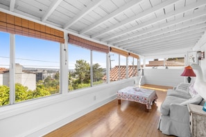 A sunroom with large windows, a wooden floor, a sofa, and a table with a floral tablecloth.