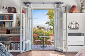 An open glass door leading to a balcony with a table and chairs, surrounded by bookshelves and decorative items.