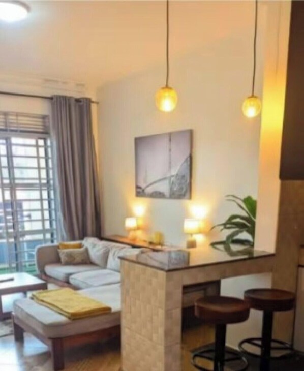 A modern living room with a sofa, a coffee table, a bar area with stools, pendant lighting, and a large window with curtains. - sereneville apartment (Kampala)