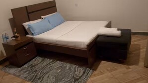 Ein ordentlich bezogenes Bett mit Kopfteil, ein Nachttisch mit Schubladen und eine kleine Bank am Fußende.
