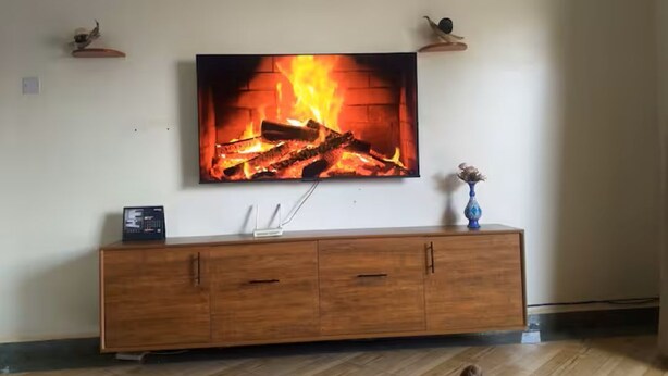 Una sala de estar con un televisor de pantalla plana montado en la pared, un mueble de madera debajo y un jarrón sobre el mueble.