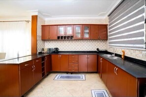 A kitchen with wooden cabinets, a black countertop, a tiled backsplash, and a window with blinds. - Evalyn apartments (Kampala)