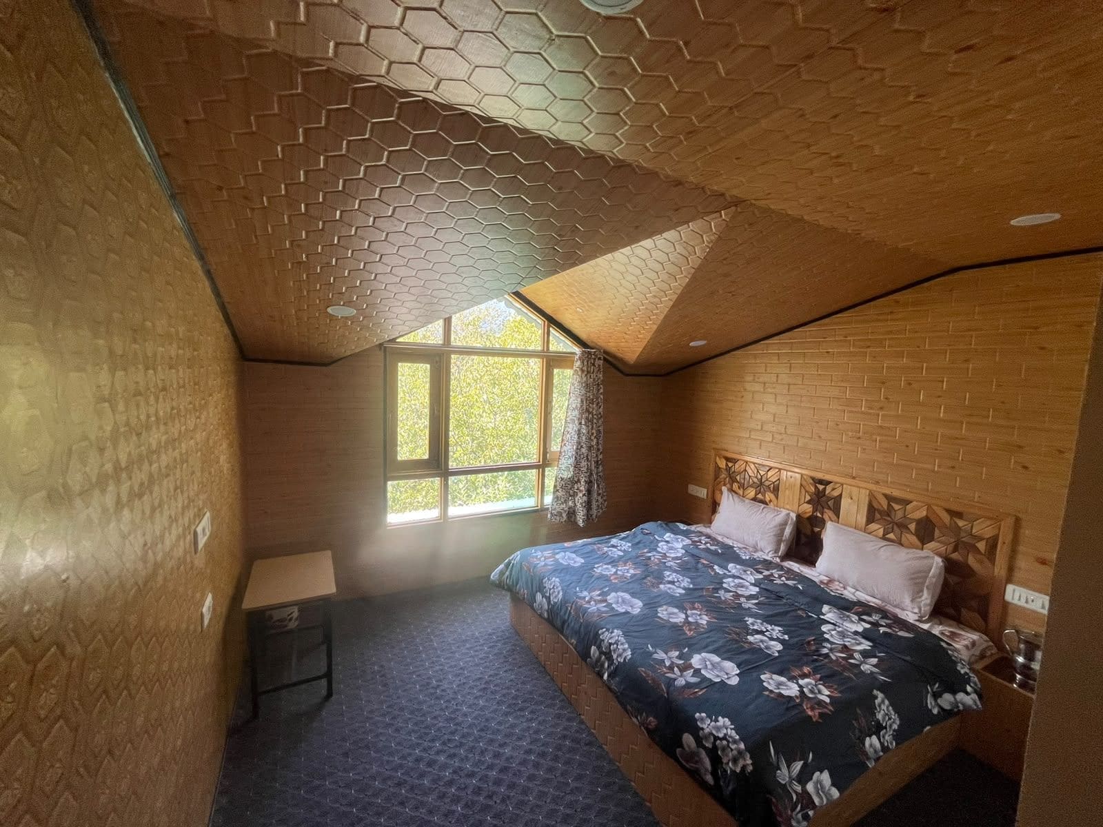 A bedroom with a brick wall, a wooden ceiling with a honeycomb pattern, a large window, a bed with floral bedding, and a small bedside table.