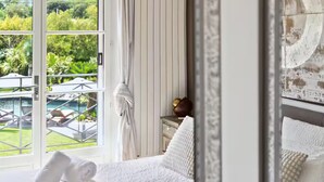Et hotelværelse med seng, et natbord, en lampe og udsigt til pool og grøn bevoksning.