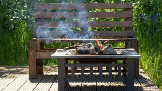 A wooden bench with a fire burning on it, surrounded by greenery.