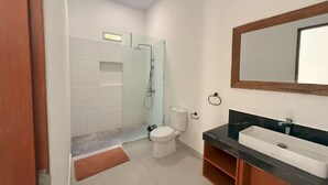 Un baño con inodoro blanco, cabina de ducha con mampara de cristal, encimera negra con lavabo y mueble de tocador de madera.