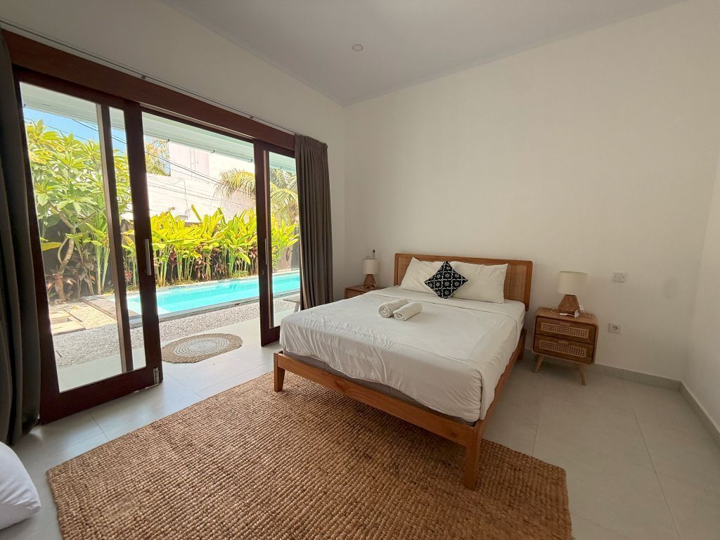 A bedroom with a bed, a nightstand, a lamp, and a view of a swimming pool.