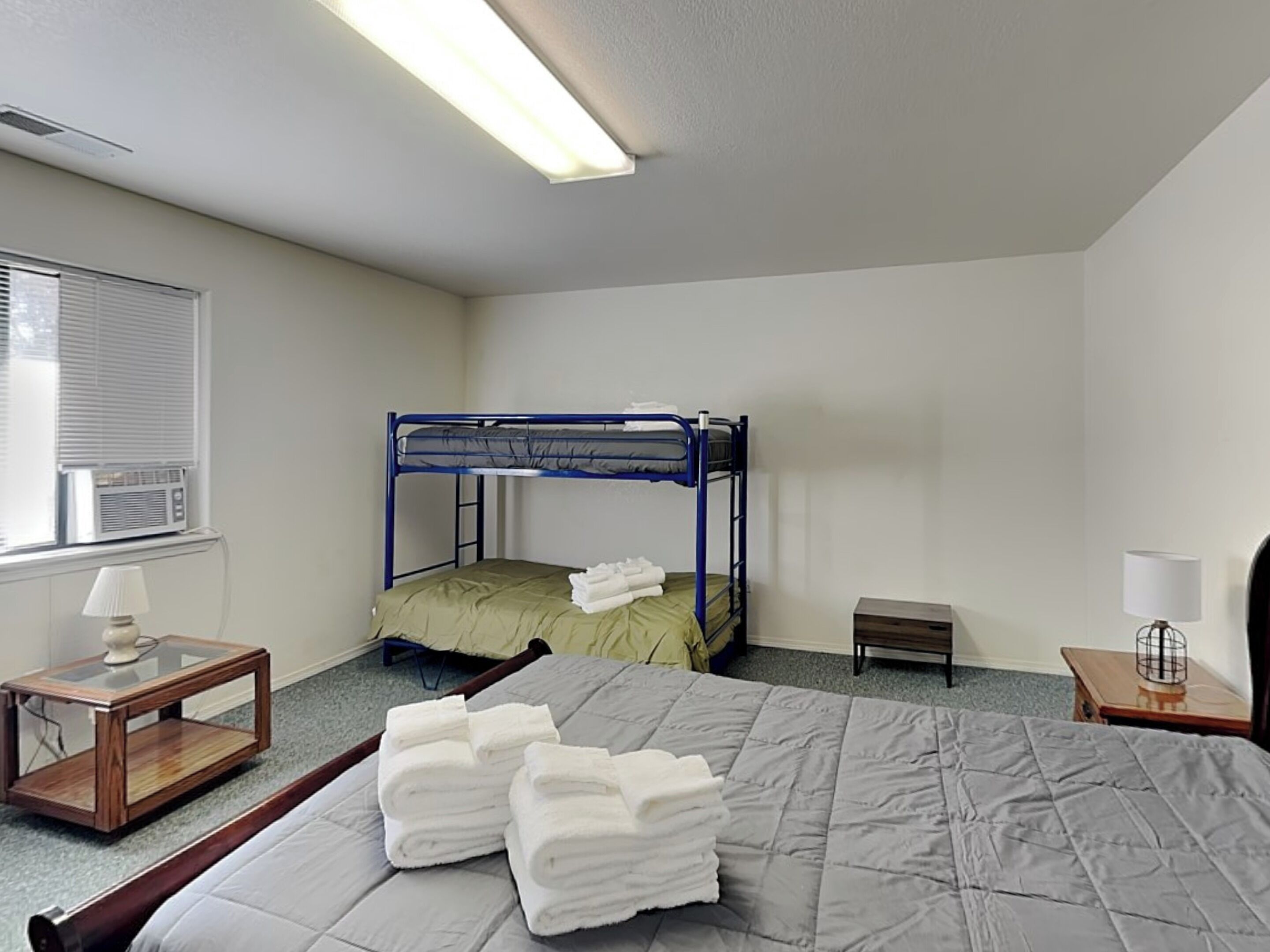 A single-bed room with a bunk bed, a bedside table with a lamp, a small bedside table with a lamp, and a window with blinds.