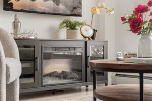 Una sala de estar moderna con chimenea, un televisor montado en la pared, una planta en maceta, un reloj y un florero con flores.