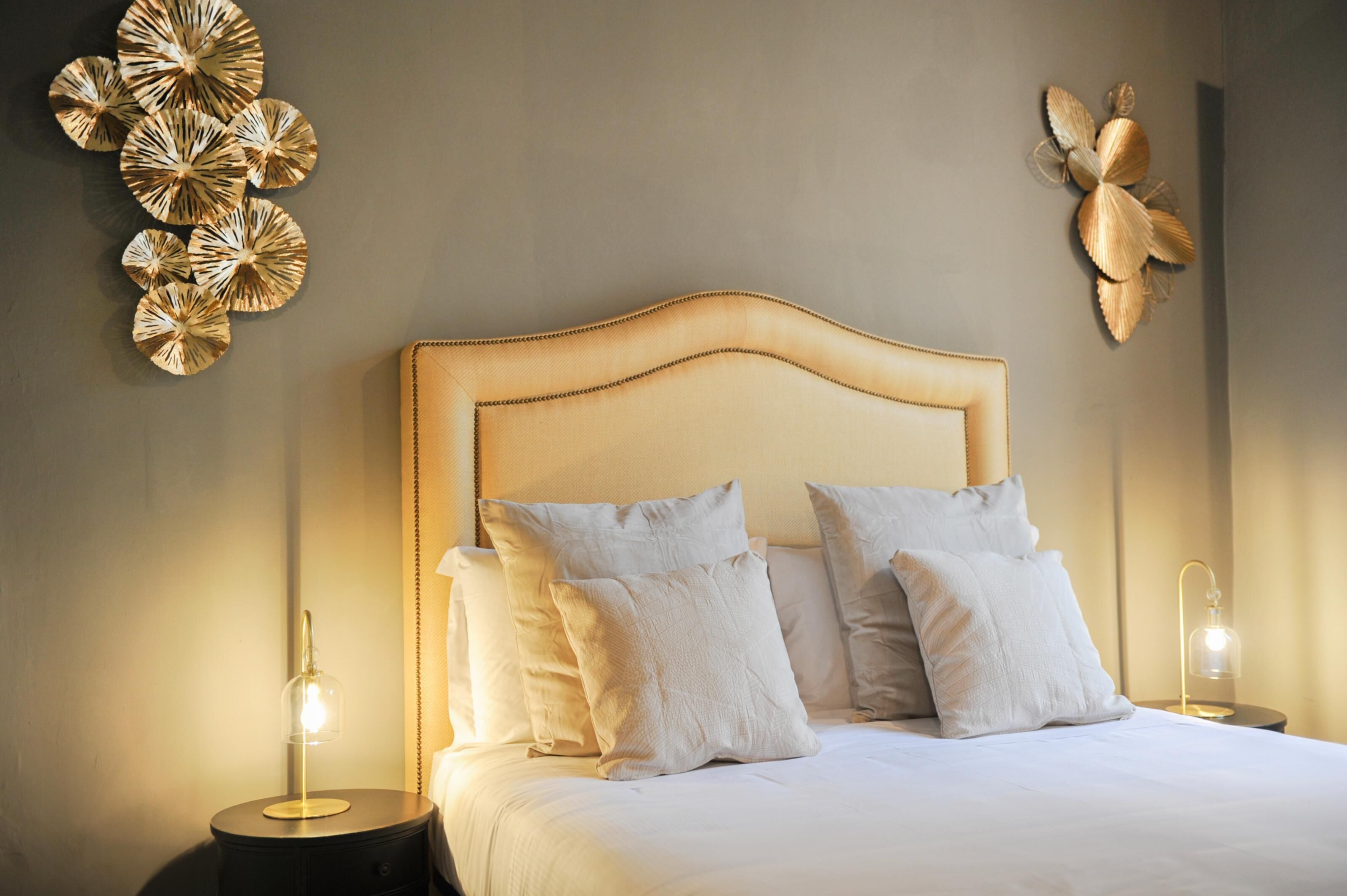 Una cama bien hecha con ropa blanca y almohadas, una mesita de noche redonda con una lámpara, y decoración en la pared.