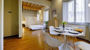 Ein Zimmer mit Holzboden, weißen Möbeln, einem gedeckten Esstisch mit Tellern und Gläsern sowie einem Fenster mit Vorhängen.