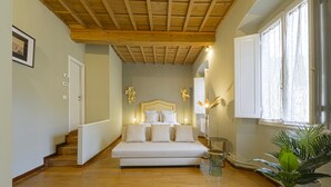 Ein Schlafzimmer mit Holzboden, einem Bett mit weißer Bettwäsche, einem Nachttisch, einer Lampe und einem Fenster mit Vorhängen.