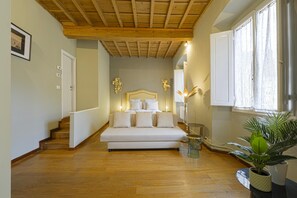 Ein Schlafzimmer mit Holzboden, einem Bett mit weißer Bettwäsche, einem Nachttisch, einer Lampe und einem Fenster mit Vorhängen.