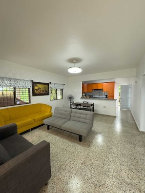 A living room with a sofa, a grey couch, a dining table, and a kitchen area with wooden cabinets. - Brisa del Rio Jimenoa (Jarabacoa)