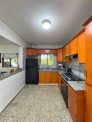 A kitchen with granite countertops, wooden cabinets, a black refrigerator, a stove, and a window with a view of greenery. - Brisa del Rio Jimenoa (Jarabacoa)