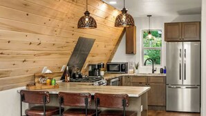 A kitchen with a wooden sloped ceiling, a stainless steel refrigerator, a microwave, and a bar area with stools. - The Timber Triangle-Views, Hot tub, Firepit (Florissant)