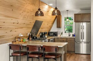 A kitchen with a wooden sloped ceiling, a stainless steel refrigerator, a microwave, and a bar area with stools. - The Timber Triangle-Views, Hot tub, Firepit (Florissant)