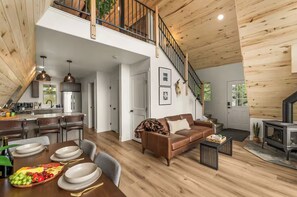 Un vaste espace de vie doté d’un haut plafond, d’un escalier, d’une cheminée et d’un coin repas avec une table et des chaises en bois.