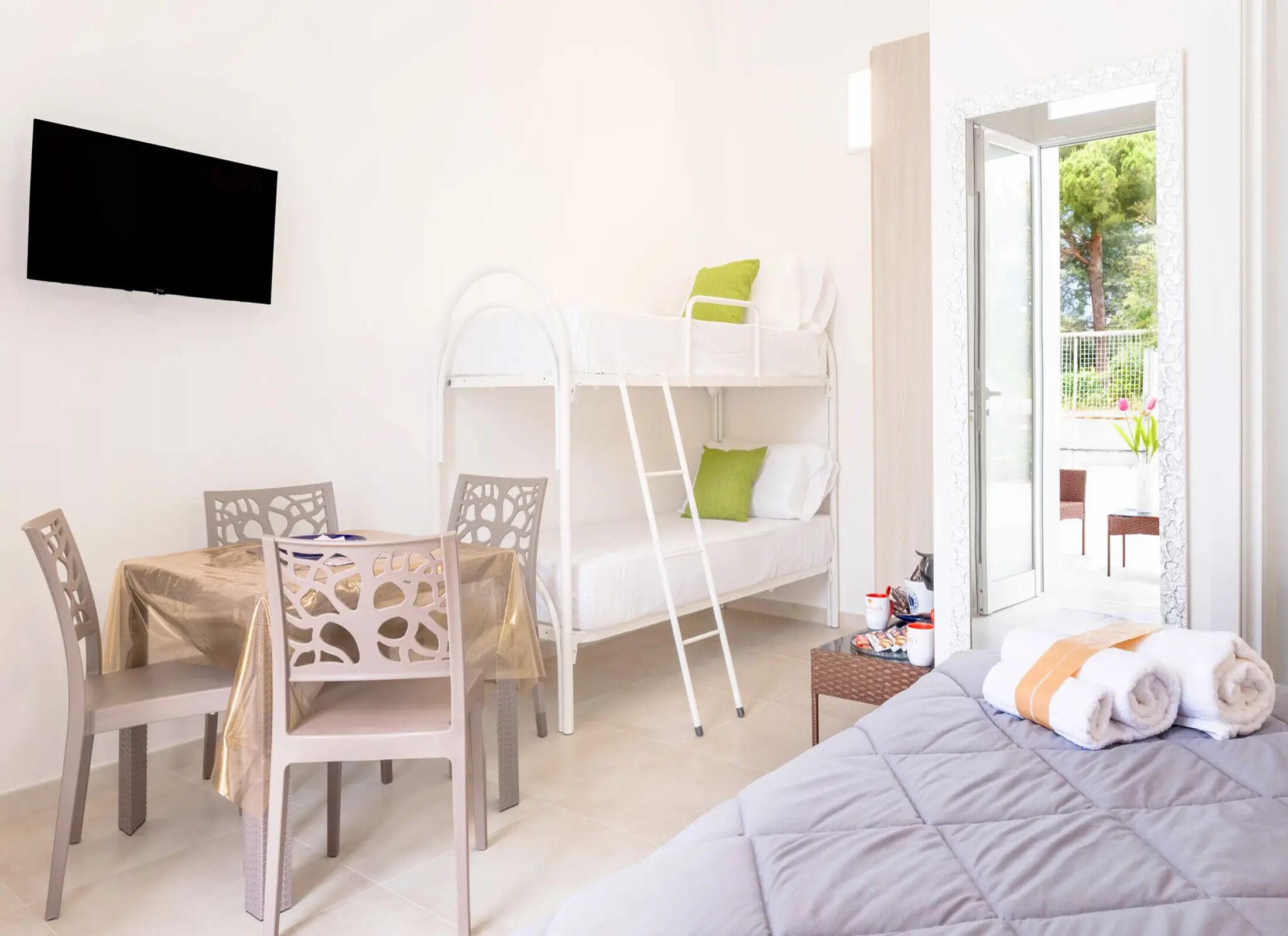 Una habitación con literas, una mesa con sillas, un televisor montado en la pared y un balcón con mobiliario exterior.