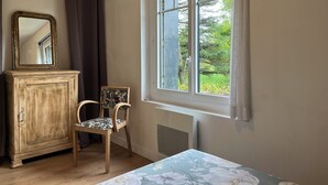 A room with a wooden cabinet, a chair, a bed with a floral bedspread, and a window with a view of greenery. - New 2-⭐️ cottage on a Loire island, ideal for families (Gennes-Val-de-Loire)