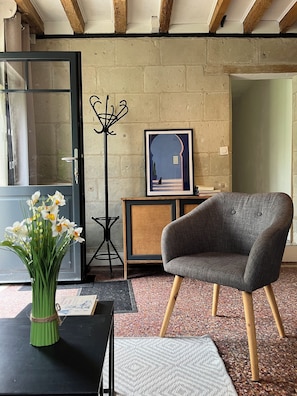 Ein Zimmer mit einem grauen Sessel, einem gerahmten Bild auf einer Holzkommode, einer Blumenvase und einem schwarzen Kleiderständer.
