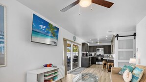 Uma sala de estar moderna com uma mesa de centro de vidro, um sofá com almofadas, um aparador branco com livros e um quadro de uma cena de praia.