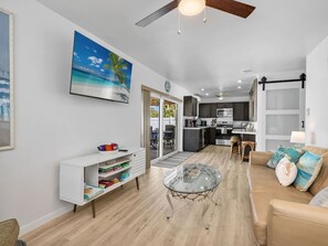 A modern living room with a glass coffee table, a sofa with cushions, a white console table with books, and a painting of a beach scene. - Pearl Beach House - Walk to FMB, Pool & Hot Tub! (Fort Myers Beach)