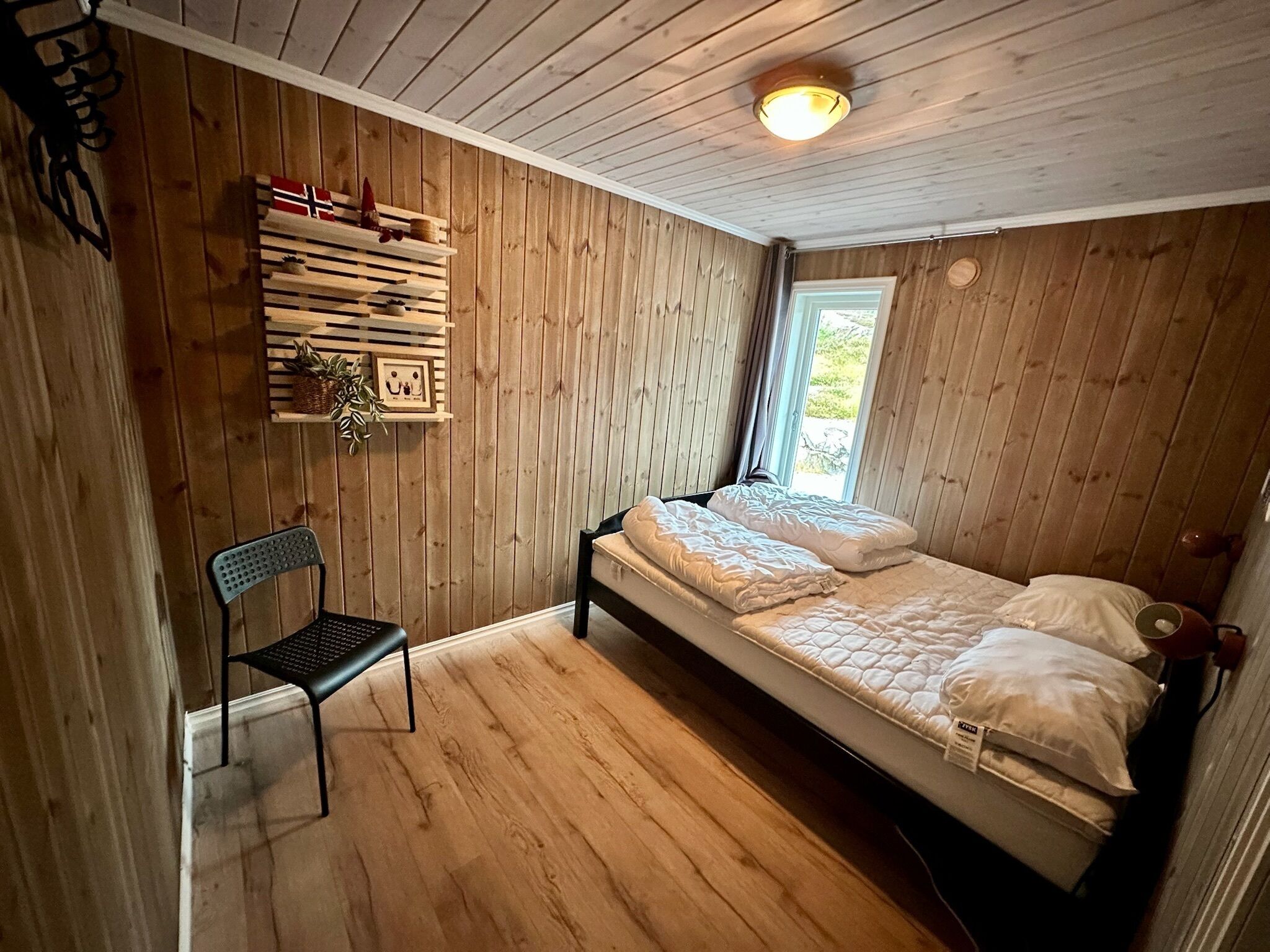 A wooden-paneled room with a bed, a chair, and a shelf with decorative items.
