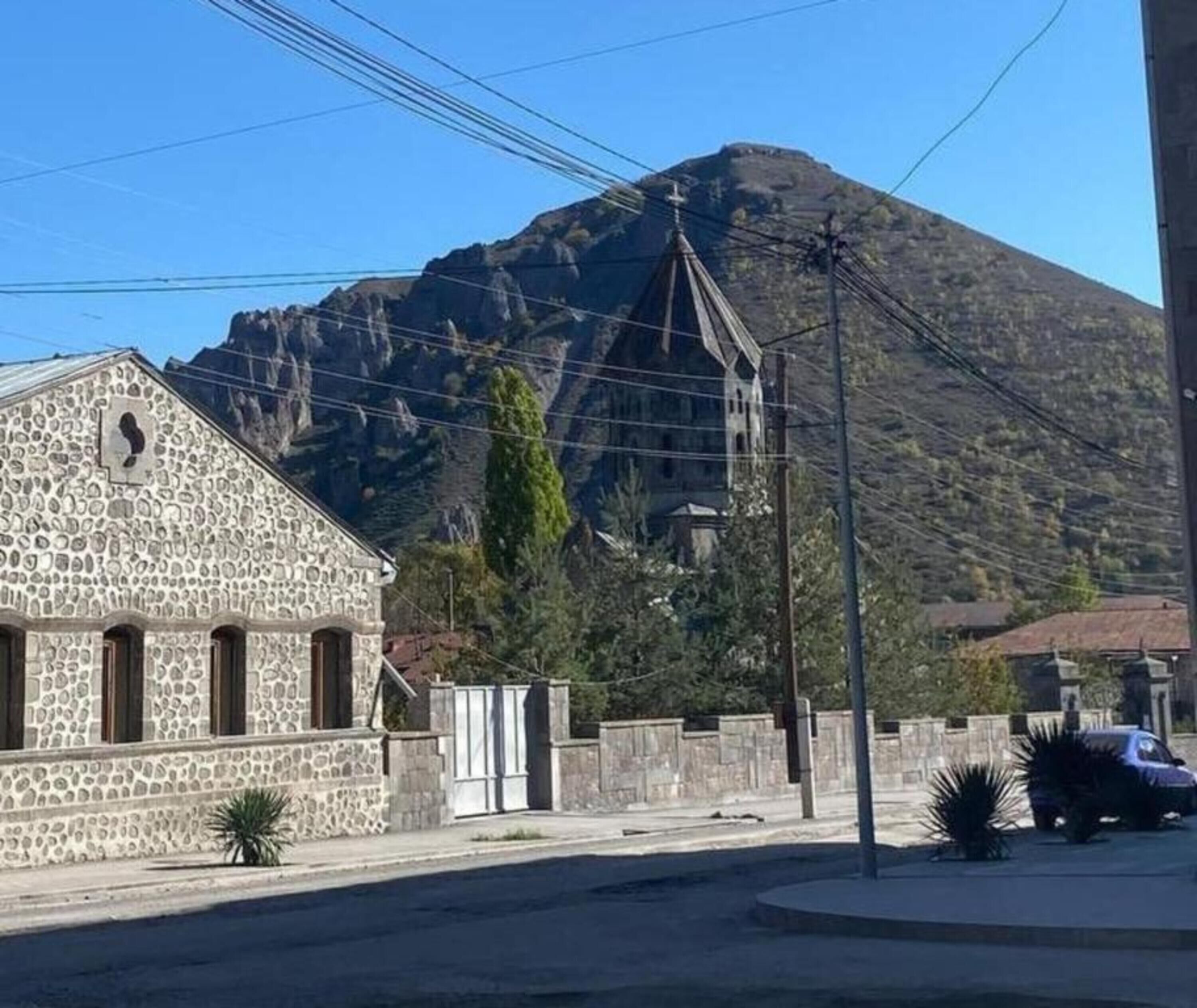 Un edificio de piedra con una cruz en la parte superior, una montaña al fondo y un coche estacionado.