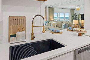 A modern kitchen with a built-in sink, a wooden cutting board with labeled bottles, and a view of the ocean through large windows. - Sandy Nook | Satellite Beach Oceanfront Condo (Satellite Beach)